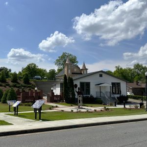 A small white building with steps, informational signs on the sidewalk, and a church with twin steeples in the background under a partly cloudy sky—Celebrate Your Event in Acworth!.