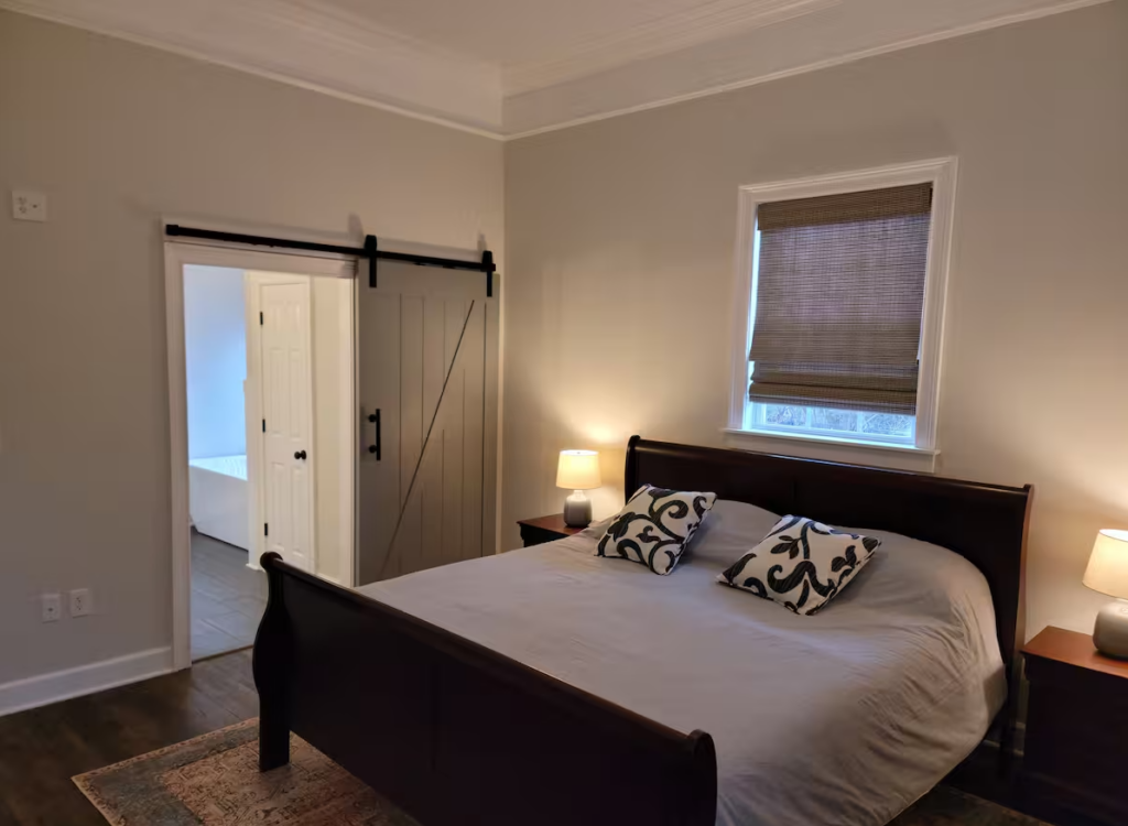 Bedroom with a wooden bed, gray bedding, two bedside tables with lamps, and a window with a brown blind. The room has a sliding barn door leading to a white-walled area.