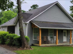 A gray house with a sloped roof, white trim, and a wooden porch featuring several rocking chairs. A tree and green shrubs border the yard.