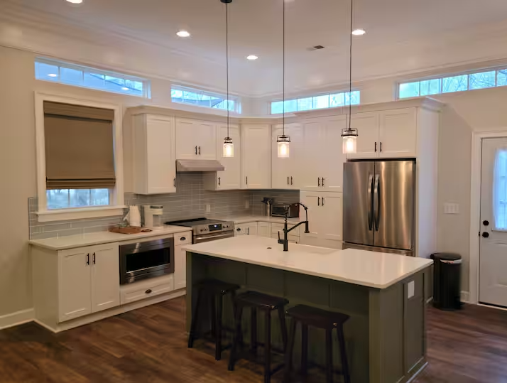 Modern kitchen with white cabinets, stainless steel appliances, and a center island with bar stools. Pendant lights hang above the island. Wood flooring and a window with a blind are visible.