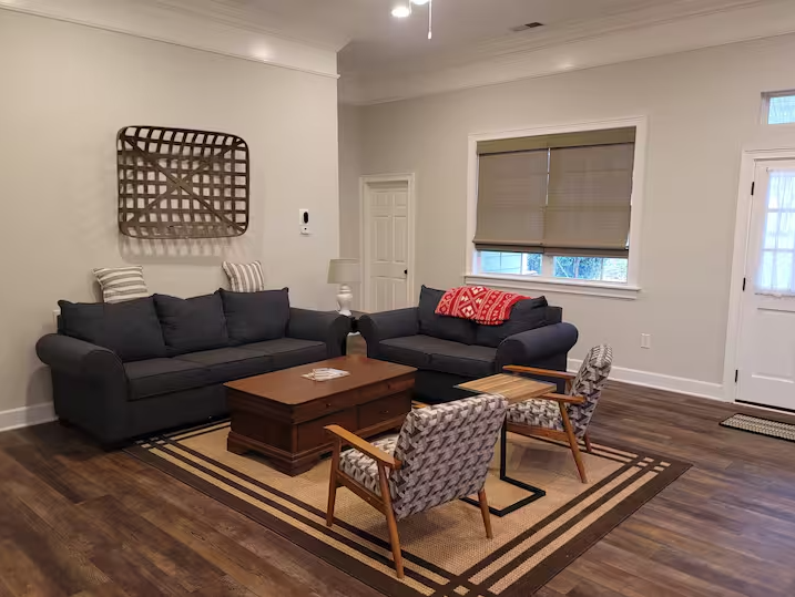 Living room with two dark sofas, wooden coffee table, two patterned chairs, wall decor, and a rug on a wooden floor. Door and window with blinds in the background.