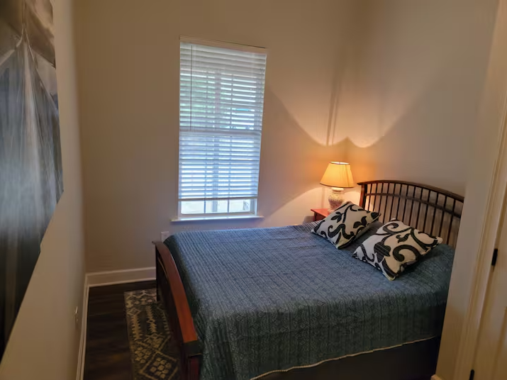 A bedroom with a wooden bed frame, blue bedspread, two patterned pillows, a bedside lamp, window with blinds, and a patterned rug on hardwood floor.