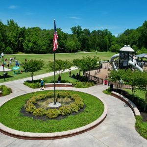 A park with a central flagpole, circular landscaping, a playground with slides, grassy fields, and people scattered throughout on a sunny day—Celebrate Your Event in Acworth!.