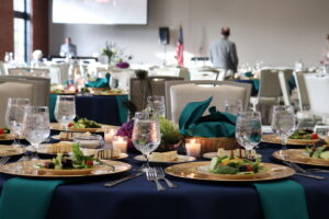 A banquet hall is set for an event, featuring round tables adorned with wine glasses, salad plates, and teal napkins. Candlelight adds a warm ambiance as foodies and fun-seekers flock to Acworth. An American flag and attendees are visible in the background.