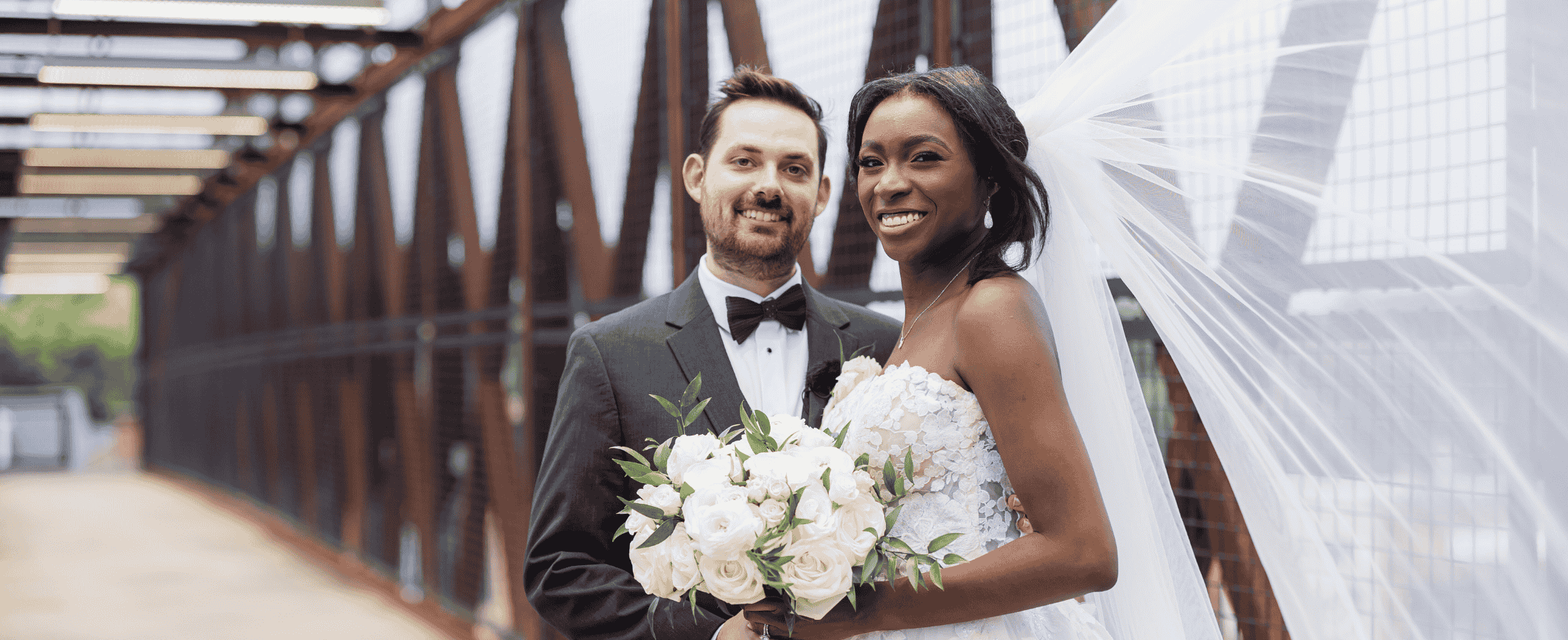 A bride and groom stand together on a bridge, holding a bouquet of white flowers. The bride wears a white dress and veil, while the groom is in a black tuxedo—a picturesque scene among many as foodies and fun-seekers flock to Acworth for romance and celebration.