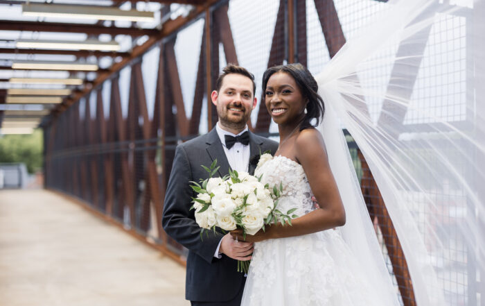 A couple in wedding attire stands on a metal bridge in Acworth, their smiles capturing the joy of the moment. The bride's veil flows gracefully as she holds a bouquet of white flowers, embodying the perfect way to celebrate your event in this picturesque location.