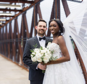 A couple in wedding attire stands on a metal bridge in Acworth, their smiles capturing the joy of the moment. The bride's veil flows gracefully as she holds a bouquet of white flowers, embodying the perfect way to celebrate your event in this picturesque location.