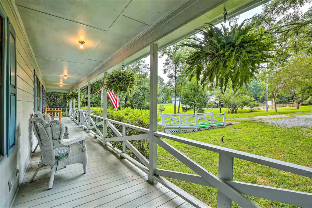 A wooden porch with white railings and wicker chairs overlooks a grassy yard. Hanging ferns and an American flag are visible. A pond and trees can be seen in the distance.