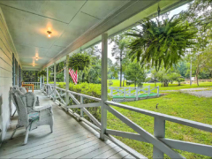 A wooden porch with white railings and wicker chairs overlooks a grassy yard. Hanging ferns and an American flag are visible. A pond and trees can be seen in the distance.