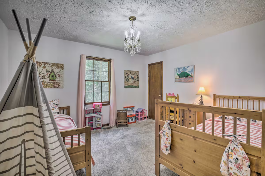 A children's bedroom with two wooden beds, a chandelier, a play tent, and wall art. A window lets in natural light, and a small lamp is on beside the door.