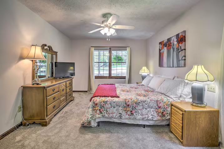 Bedroom with a floral comforter on a bed, two bedside tables with lamps, a dresser with a mirror, and a TV. Ceiling fan above and window with curtains in the background.