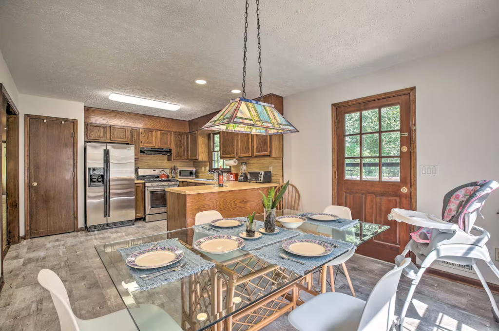 Kitchen and dining area with a glass table set for six, a high chair, a wooden door, stainless steel appliances, and wooden cabinets. Pendant light hangs above the table.