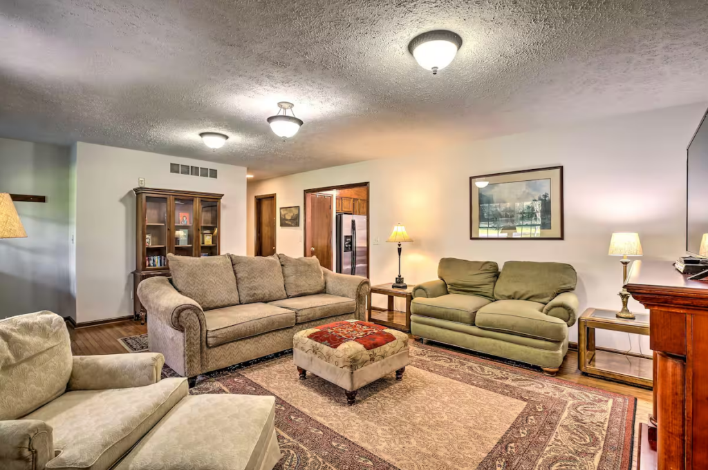 A cozy living room with two sofas, an armchair, a cushioned ottoman, wooden bookshelves, and a patterned rug. Warm lighting from lamps and ceiling fixtures. A framed picture hangs on the wall.