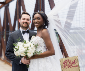 Bride and groom standing on a bridge, bride holds white flowers, groom wears a suit. Bride's veil flows behind them as they celebrate their event in Acworth. A sign in the foreground reads, "You're Welcome in Acworth.