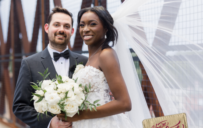 Bride and groom standing on a bridge, bride holds white flowers, groom wears a suit. Bride's veil flows behind them as they celebrate their event in Acworth. A sign in the foreground reads, "You're Welcome in Acworth.