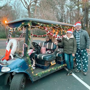 A family enjoys Holly Jolly Happenings for Everyone in Acworth, standing beside a golf cart adorned with lights, garlands, candy canes, and Santa hats. They are dressed in warm clothing, perfectly capturing the festive spirit.