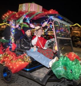 A decorated golf cart with Christmas lights, garlands, and presents, carrying two people in festive attire at night.