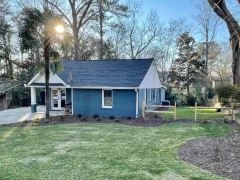 A blue house with a dark roof sits on a green lawn. A large tree is in the backyard, and the sun peeks through the branches. A wooden fence is partially visible on the right.