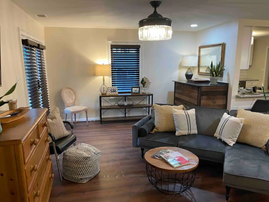 Living room with a gray sectional sofa, decorative pillows, a round wooden coffee table, a dresser, a floor lamp, a chandelier, and a small console table by the window with blinds.