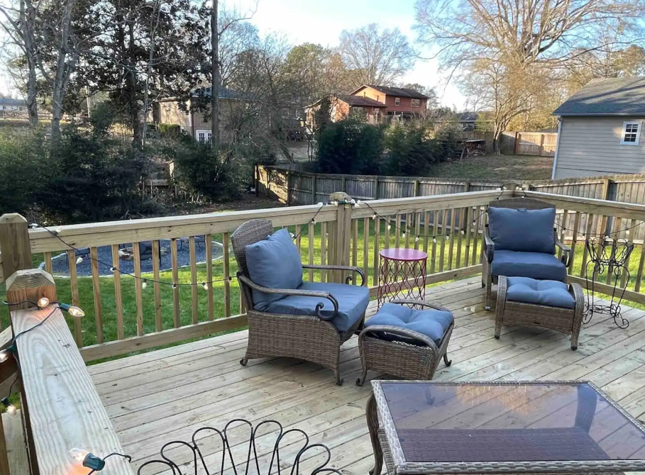 A wooden deck with wicker furniture, including two chairs with blue cushions and a round side table, overlooks a grassy backyard bordered by trees and a fence.