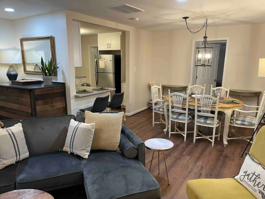 Cozy living and dining area with a gray sectional sofa, round coffee table, decorative pillows, dining table with mixed chairs, modern chandelier, and white kitchen. Wood flooring throughout.