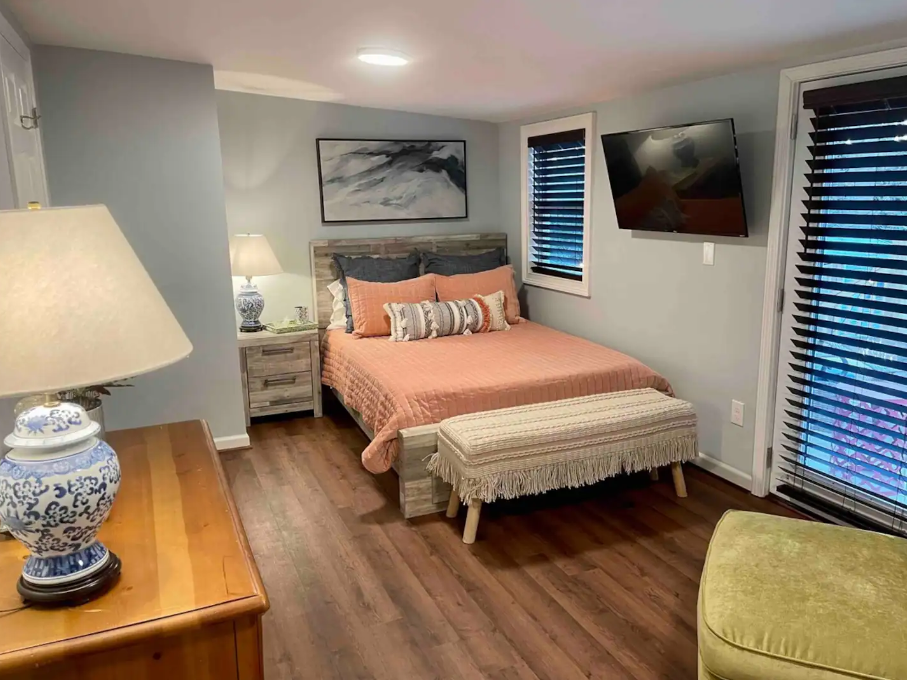 Bedroom with a bed, peach bedding, wooden headboard, and side table. Wall-mounted TV, dresser with lamp, and artwork above the bed. Two windows with blinds and a green chair are present.