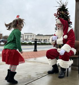 Child in festive attire approaches a seated Santa Claus outdoors near a decorated tree and buildings in the background.