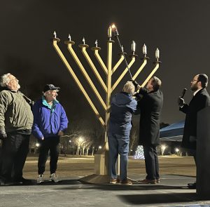 A group of people stand by a large menorah outdoors at night, with one person lighting the candles.