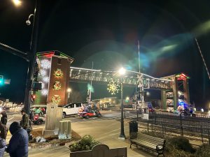 Street scene at night in Acworth, showcasing Holly Jolly Happenings. The decorated bridge twinkles with lights and wreaths as vehicles and pedestrians pass by, beneath a large snowflake light hanging elegantly.