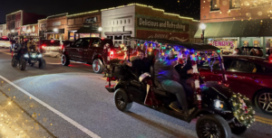 During the Holly Jolly Happenings for Everyone in Acworth, decorated vehicles adorned with lights glide down the street at night in a festive parade. People eagerly watch from sidewalks lined with illuminated shops, soaking in the cheerful holiday atmosphere.