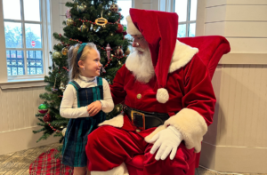 A child in a dress sits on Santa's lap amidst Holly Jolly Happenings for Everyone in Acworth, where a decorated Christmas tree adds to the festive room's charm.