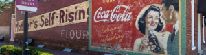 Vintage Coca-Cola mural on a brick wall featuring a woman holding a bottle, a man looking on, and an advertisement for self-rising flour.