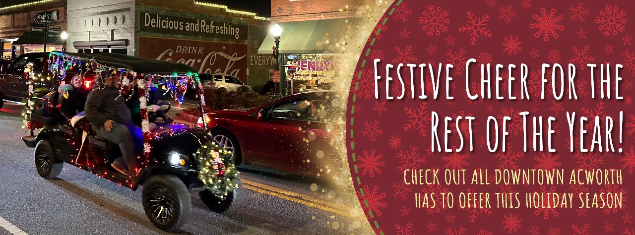 A decorated vehicle with people wearing winter clothes drives through a lit-up street. A festive sign on the right promotes holiday events in downtown Acworth.