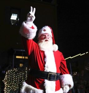 Santa Claus standing outside at night, raising his hand in a peace sign gesture, with bright festive lights in the background.
