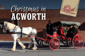 A horse-drawn carriage with passengers in festive attire rides on a street in front of a brick building. Sign reads "Christmas in Acworth" with a small banner saying "You're Welcome in Acworth.