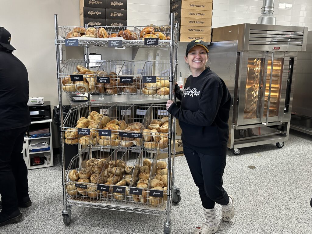 Person standing next to a rack filled with various bagels in front of an industrial oven in a bakery setting.