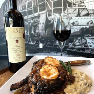 A plated steak with sauce and goat cheese sits on mashed potatoes with green beans, next to a bottle and glass of red wine—one of Acworth's must-visit spots for the perfect Valentine's Day. A black and white train mural is in the background.