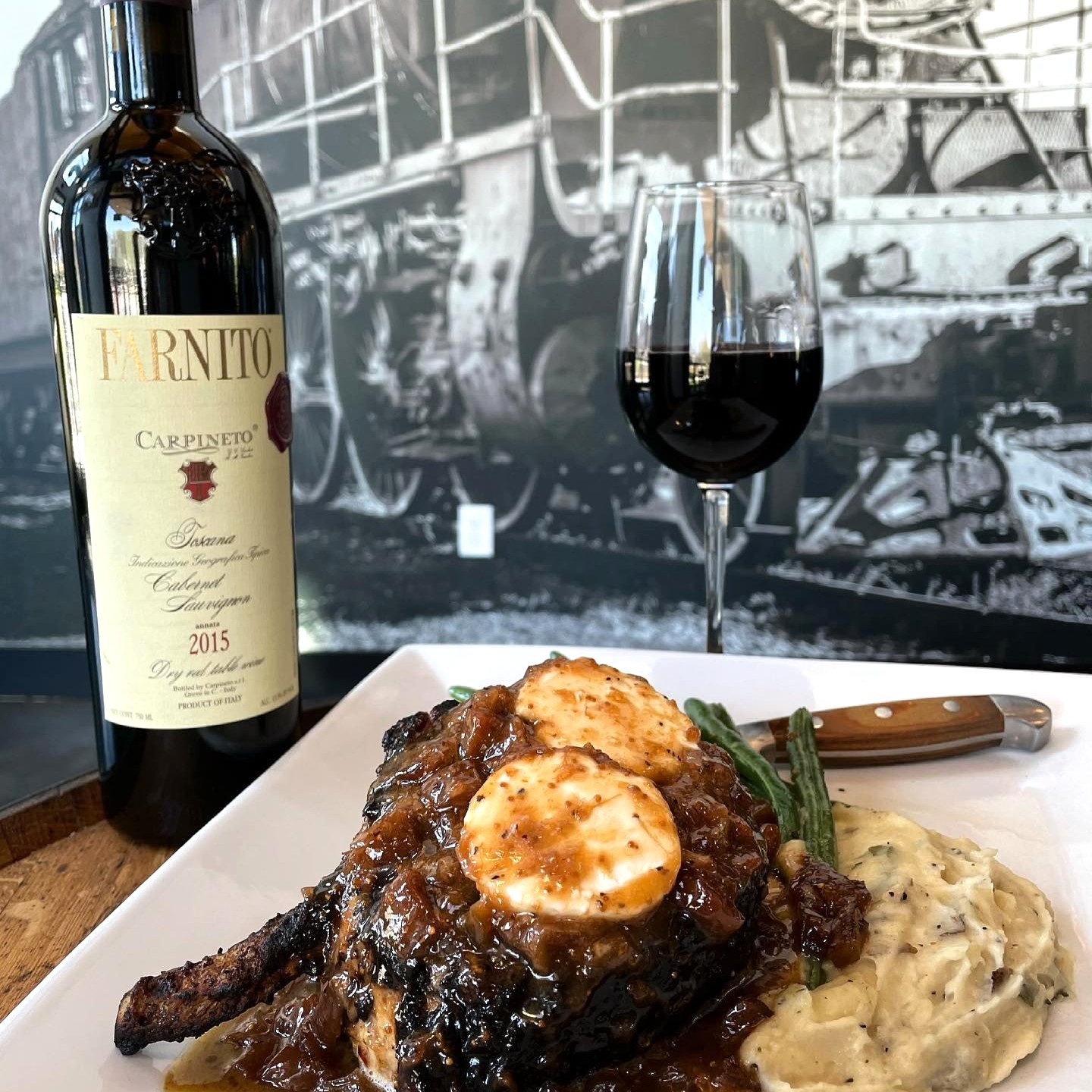 A plated steak with sauce and goat cheese sits on mashed potatoes with green beans, next to a bottle and glass of red wine—one of Acworth's must-visit spots for the perfect Valentine's Day. A black and white train mural is in the background.