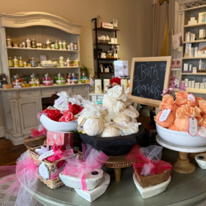 A cozy boutique, one of Acworth's 6 must-visit spots for the perfect Valentine's Day, displays bath bombs in bowls on a circular table. Shelves featuring various skincare products fill the background, complemented by a small chalkboard sign that reads "Bath Bombs.