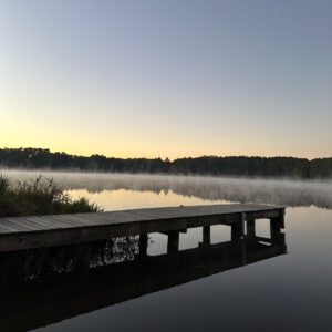 A wooden dock extends over a calm lake with mist rising, surrounded by trees under a clear sky and a faint trail from an aircraft—one of the 5 must-visit Acworth spots for Valentine's Day.