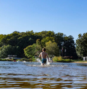 A boy runs into a lake with trees in the background and a clear blue sky, capturing one of five new reasons to put Acworth on your travel bucket list. Meanwhile, two adults and two children stand on the sandy shore to the right, soaking in the scenic beauty.