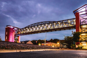 Elevated walkway labeled "Kenworth" spans a road, supported by red brick pillars, with streetlights and buildings in the background under a cloudy sky.