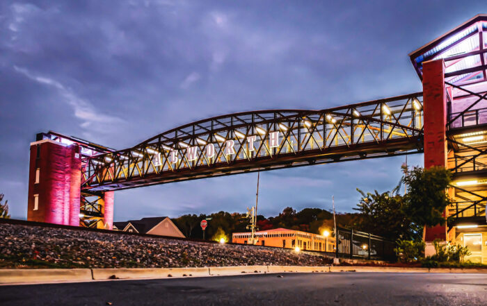 Elevated walkway labeled "Kenworth" spans a road, supported by red brick pillars, with streetlights and buildings in the background under a cloudy sky.