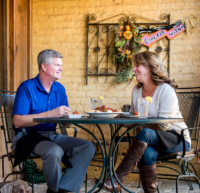 A man and woman sit at an outdoor cafe table, enjoying a delightful meal. Their small dog rests peacefully under the table. Decorative wall art adorns the background, capturing the charm of one of Acworth's 6 must-visit spots for the perfect Valentine's Day.