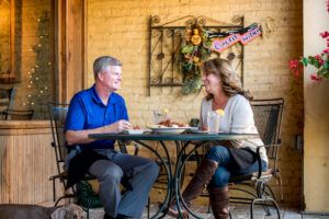 A man and woman sit at an outdoor cafe table, enjoying a delightful meal. Their small dog rests peacefully under the table. Decorative wall art adorns the background, capturing the charm of one of Acworth's 6 must-visit spots for the perfect Valentine's Day.