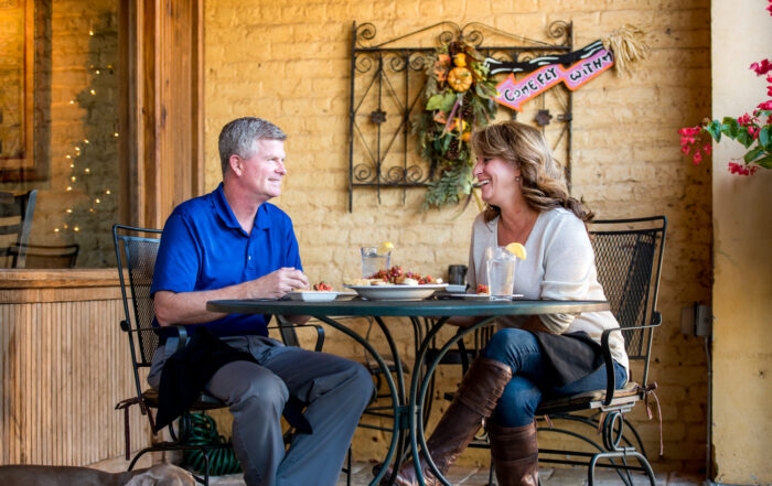 A man and woman sit at an outdoor cafe table, enjoying a delightful meal. Their small dog rests peacefully under the table. Decorative wall art adorns the background, capturing the charm of one of Acworth's 6 must-visit spots for the perfect Valentine's Day.