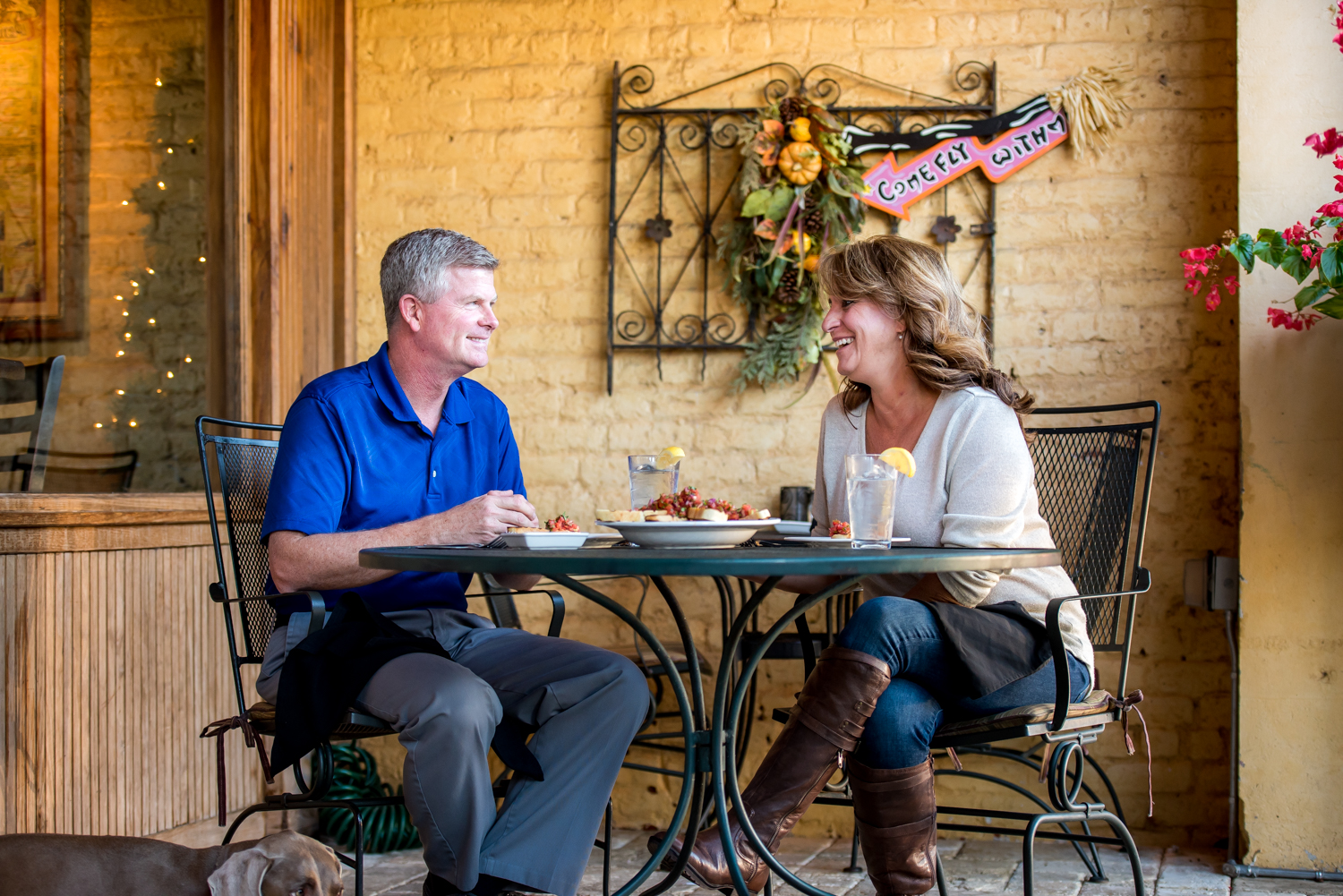 A man and woman sit at an outdoor cafe table, enjoying a delightful meal. Their small dog rests peacefully under the table. Decorative wall art adorns the background, capturing the charm of one of Acworth's 6 must-visit spots for the perfect Valentine's Day.