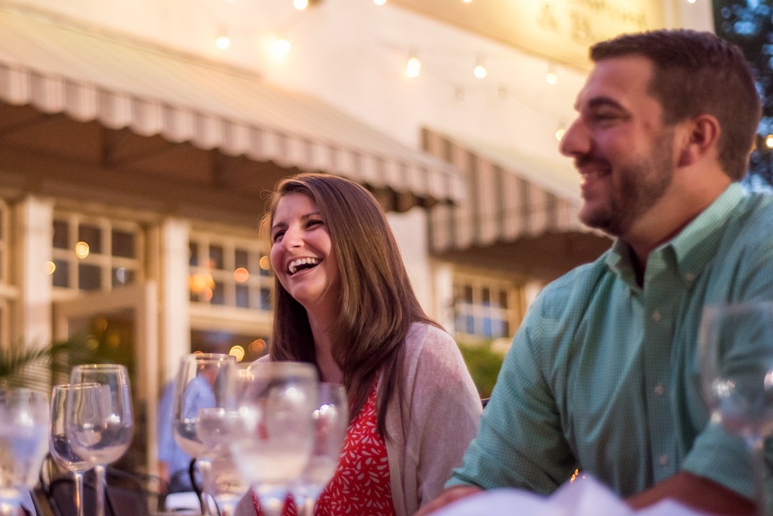Two people sit at an outdoor restaurant table, smiling and laughing under string lights. Discover moments like this at one of the 6 MUST-VISIT SPOTS FOR THE PERFECT VALENTINES DAY IN ACWORTH.