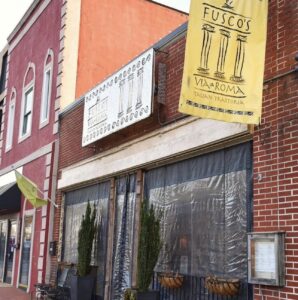 The exterior of Fusco's Via Roma Italian Trattoria, with its charming yellow sign and brick facade, is just one of five new reasons to put Acworth on your travel bucket list. Nearby, the street buzzes with life and additional storefronts await exploration.