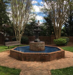 Among the 5 must-visit Acworth spots for Valentine's Day, this charming brick fountain features water in an octagonal basin, surrounded by trees and a wooden bench, set in a grassy area with a clear blue sky above.